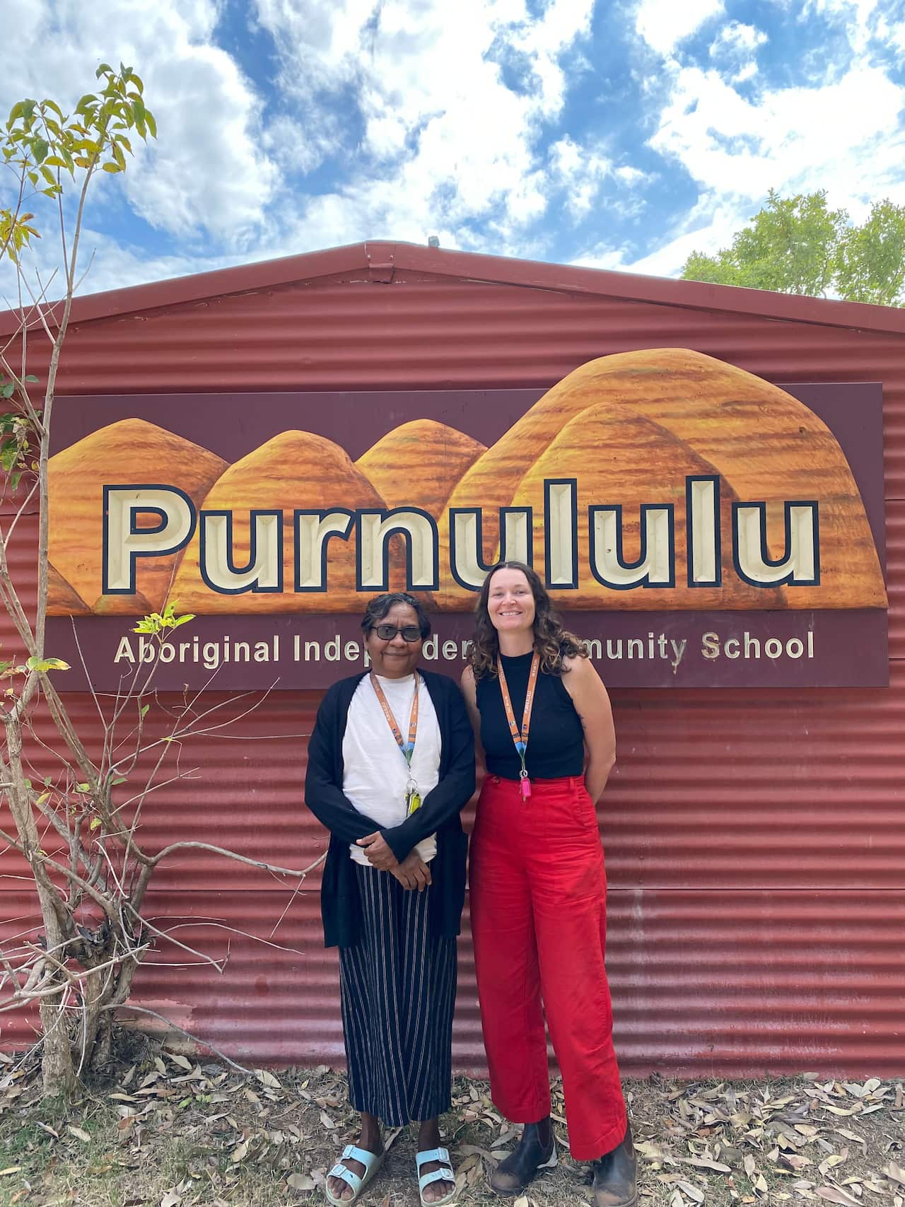 Two people smiling in front of sign that reads Purnululu in large letters, with Aboriginal Independent Community School written below it. The sign is on a red-coloured building