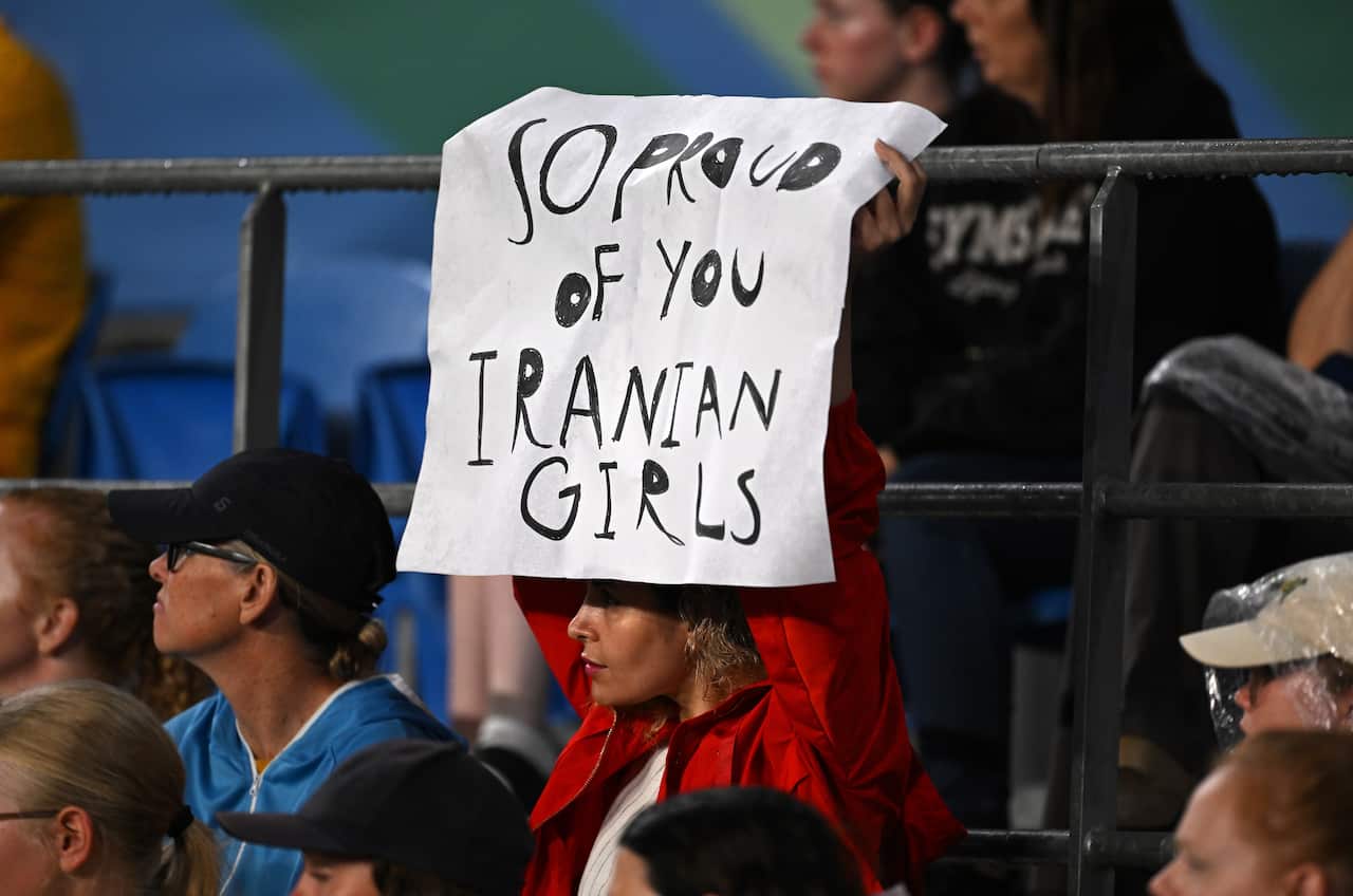 A woman in a red jacket holding up a sign that says "So proud of you Iranian girls".