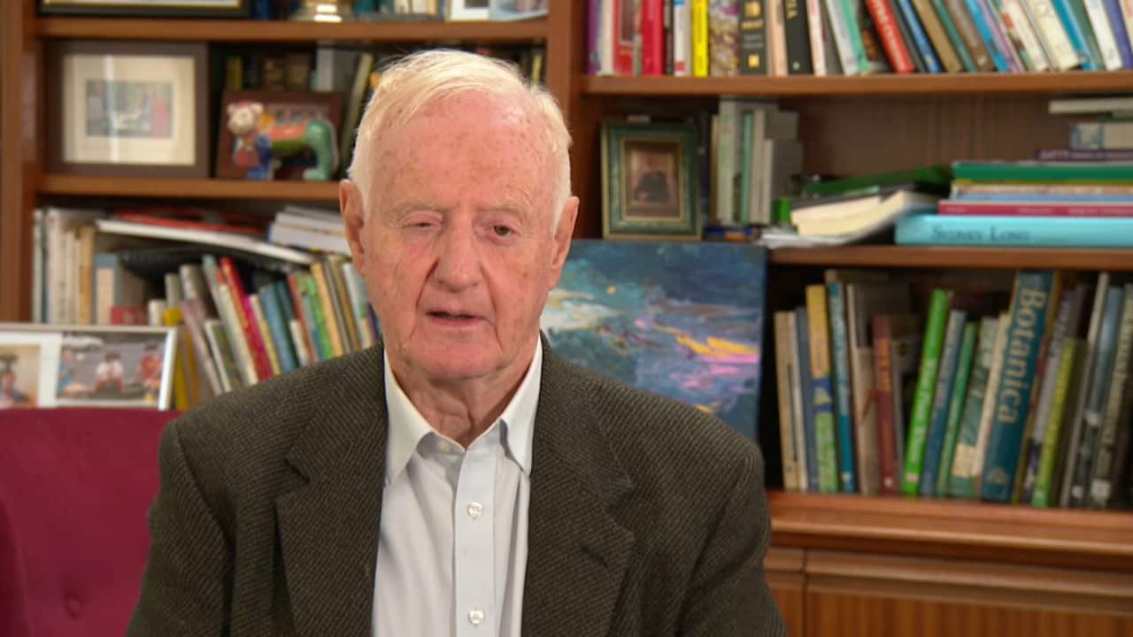 A man in a grey suit is interviewed while he is seated in front of a bookshelf. 