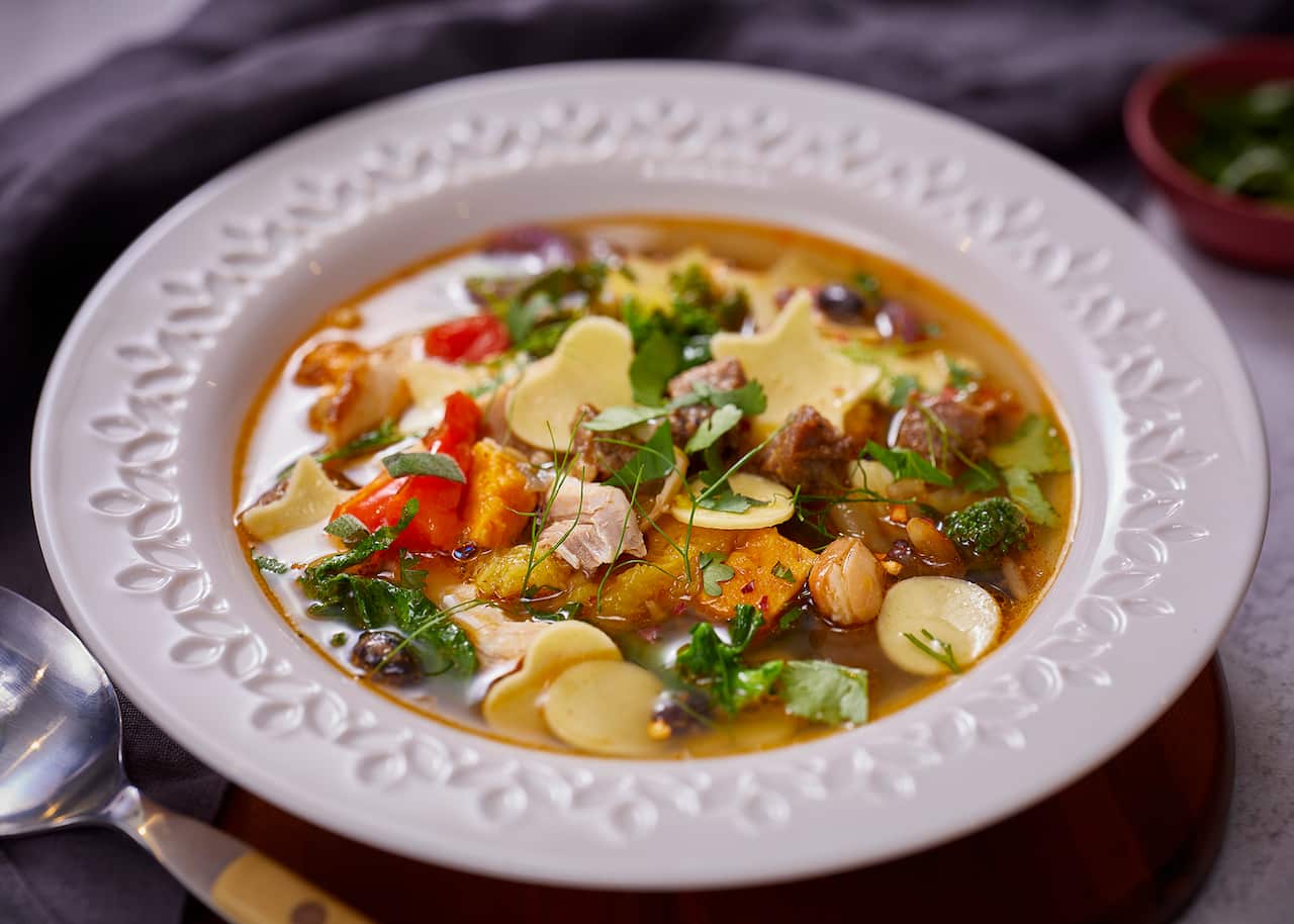 A wide white soup bowl with a textured floral pattern on the rim holds a birght soup, full of vegetables, meat and herbs. 