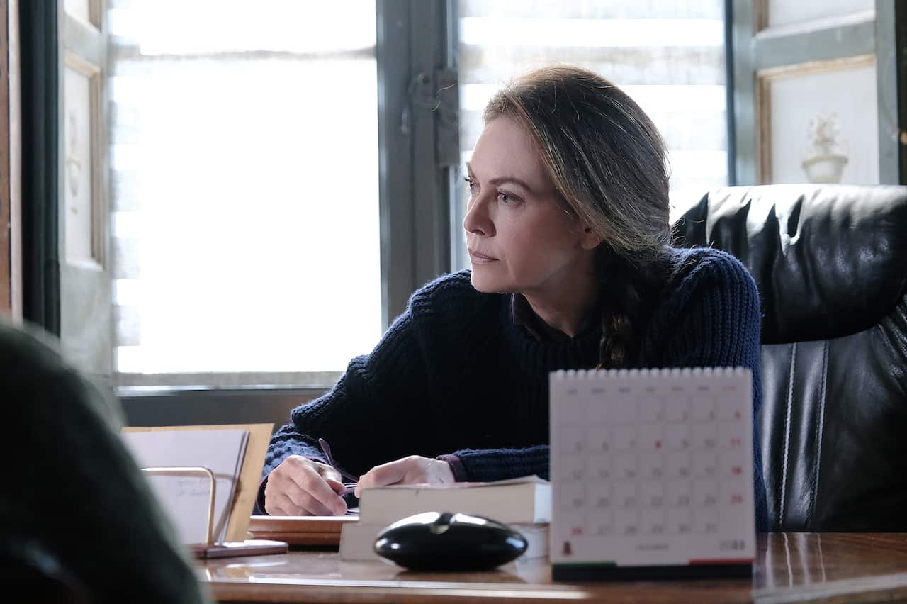 A woman sits at a desk in front of a windown. She looks pensively to one side.