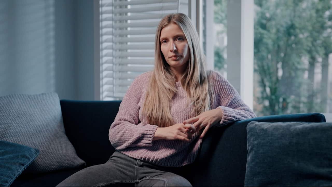 A woman with long straight blonde hair wearing grey jeans and a pastel pink sweater seated on a sofa with her hands interlaced