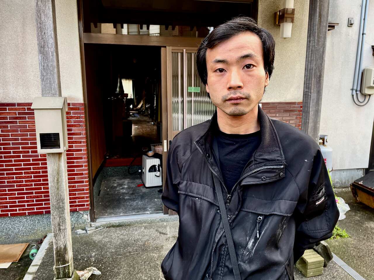 A man with a sad expression on his face wearing a black jacket stands in front of the entrance to a house. 