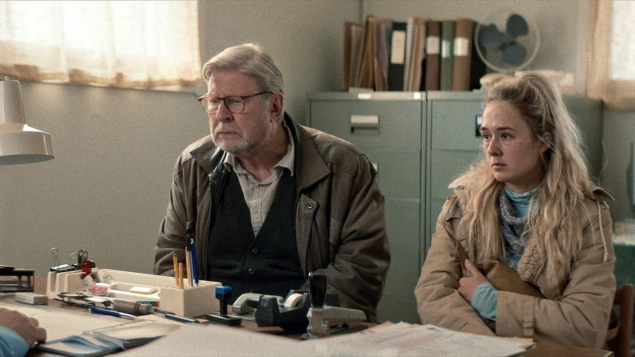 An older man and a young woman sit at a desk in an office, looking tense. 