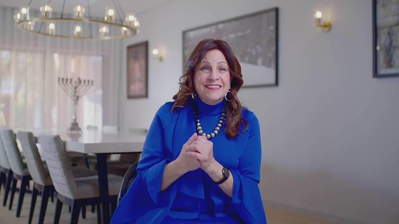 A woman in a blue dress sits in a home dining room, her hands are clasped and she is smiling. 