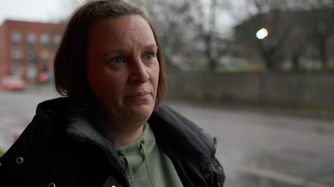 A woman with short hair and a black puffer jacket stands outside. Behind her is a park and a row of buildings, out of focus. She has a neutral facial expression. 