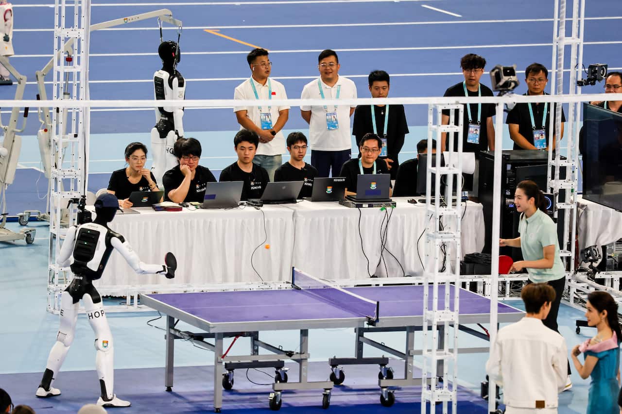 A robot and a human play table tennis surrounded by spectators on laptops.