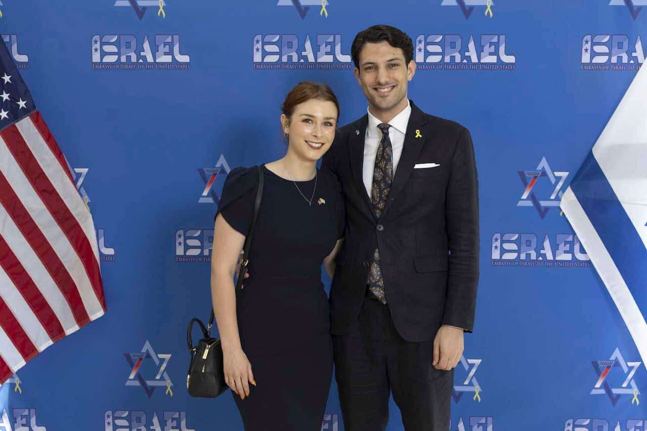 A woman and a man in black formal outfits are standing in front of a blue background.