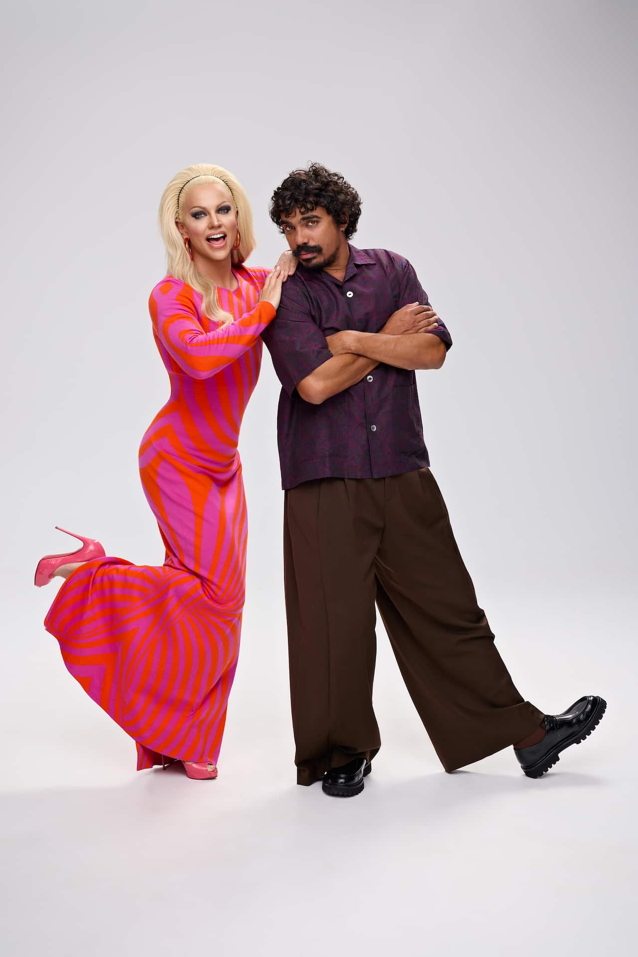 A man in a black outfit stands with his hands folded, posing next to a woman with long blonde hair and wearing a red dress. Her hands are resting on his shoulder.