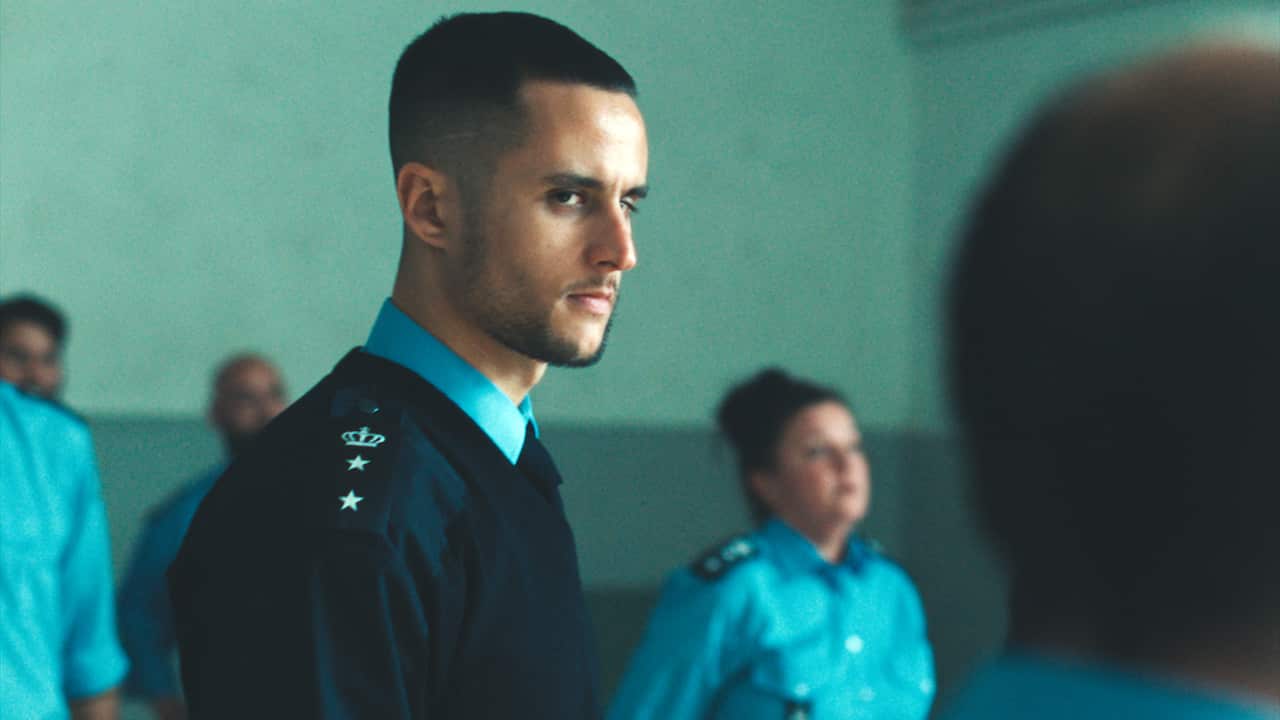 A young man in a prison guard uniform stands in a room with grey-blue walls. Several other staff in uniform can be seen behind him. 