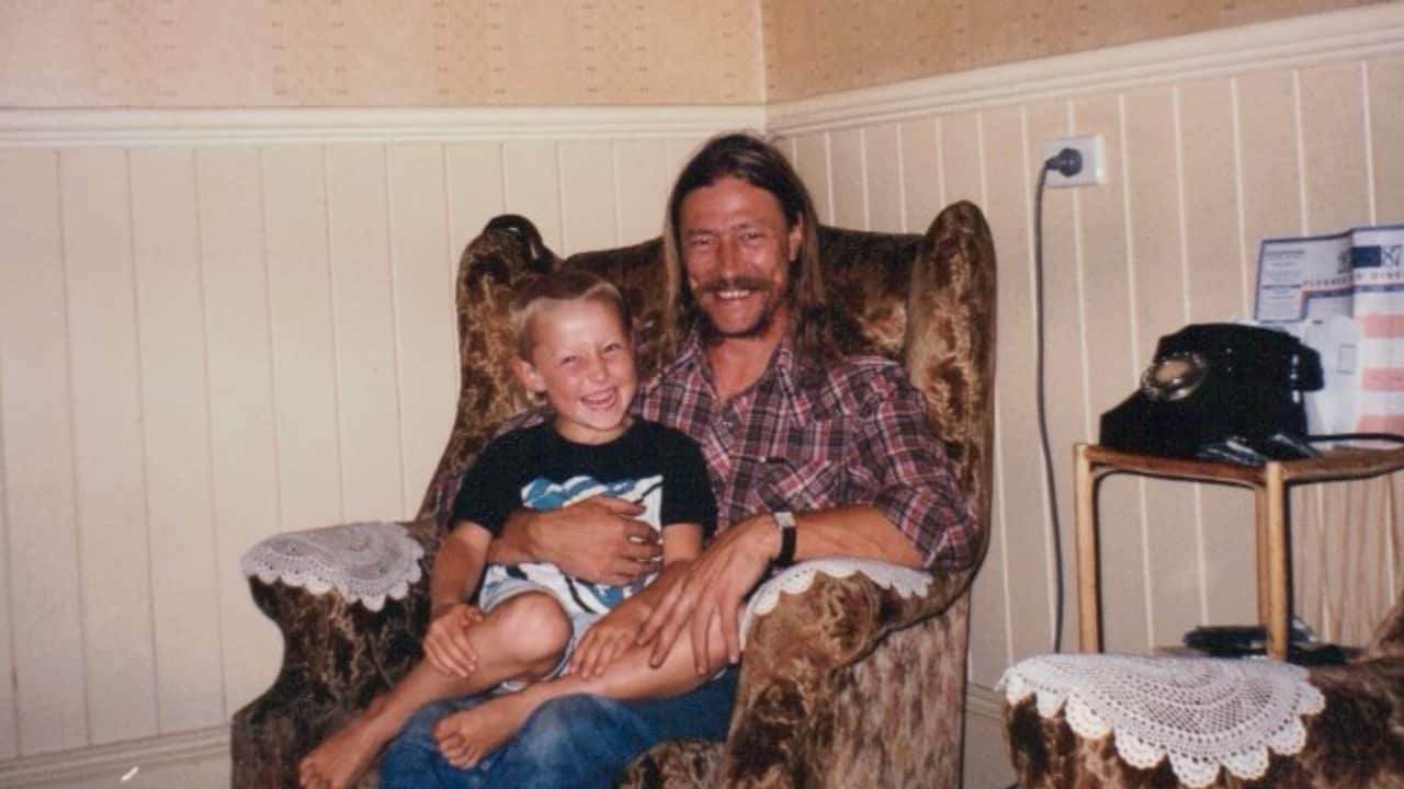 A middle-aged blonde man with a beard sits on an armchair in a living room with a young smiling boy on his lap
