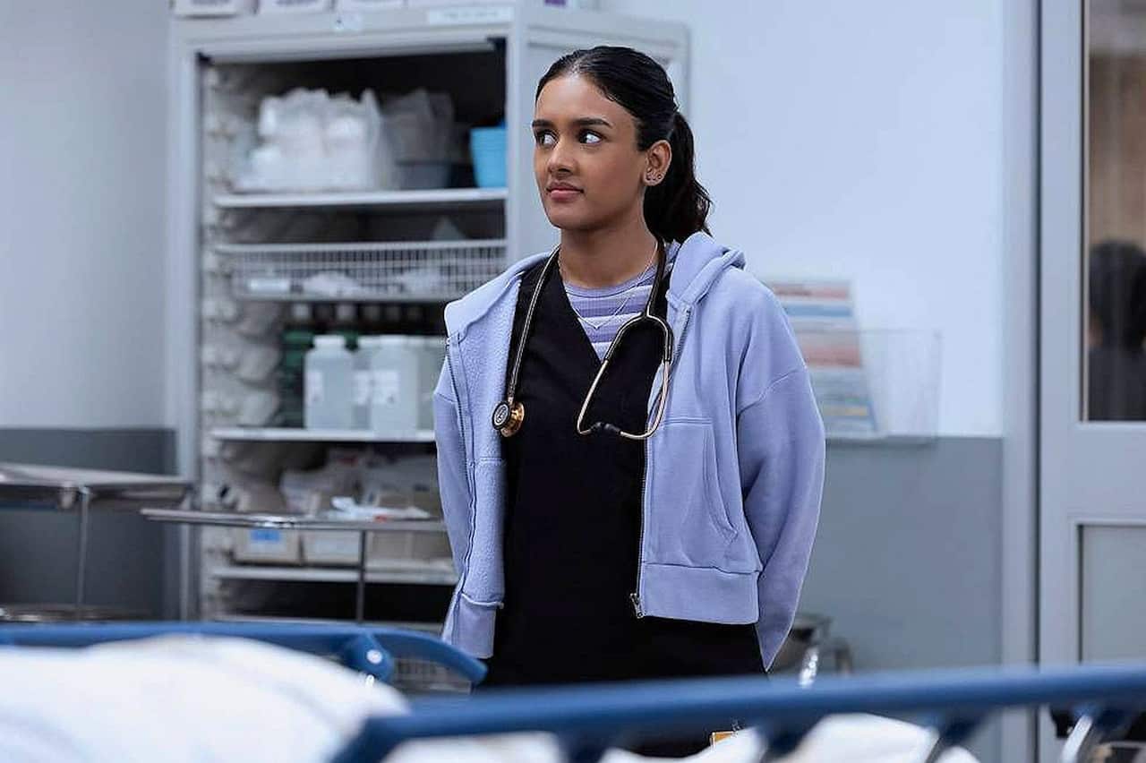 A female doctor in black scrubs and a mauve jacket with a stethoscope around her neck stands in a hospital room.