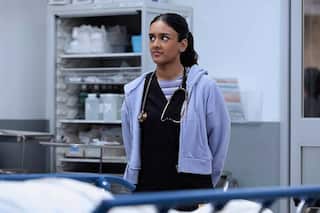 A female doctor in black scrubs and a mauve jacket with a stethoscope around her neck stands in a hospital room.