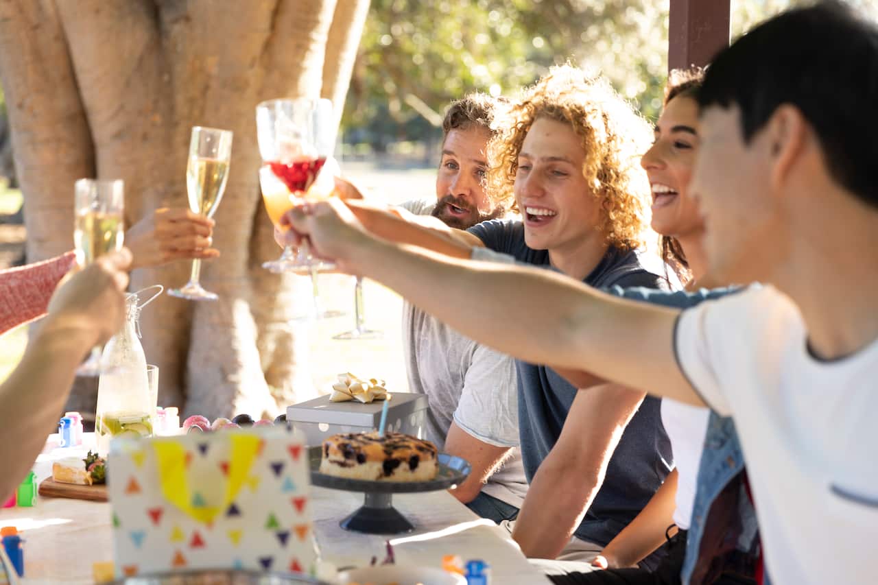 Group of young people sitting at a table outside and raising glasses for a birthday toast 