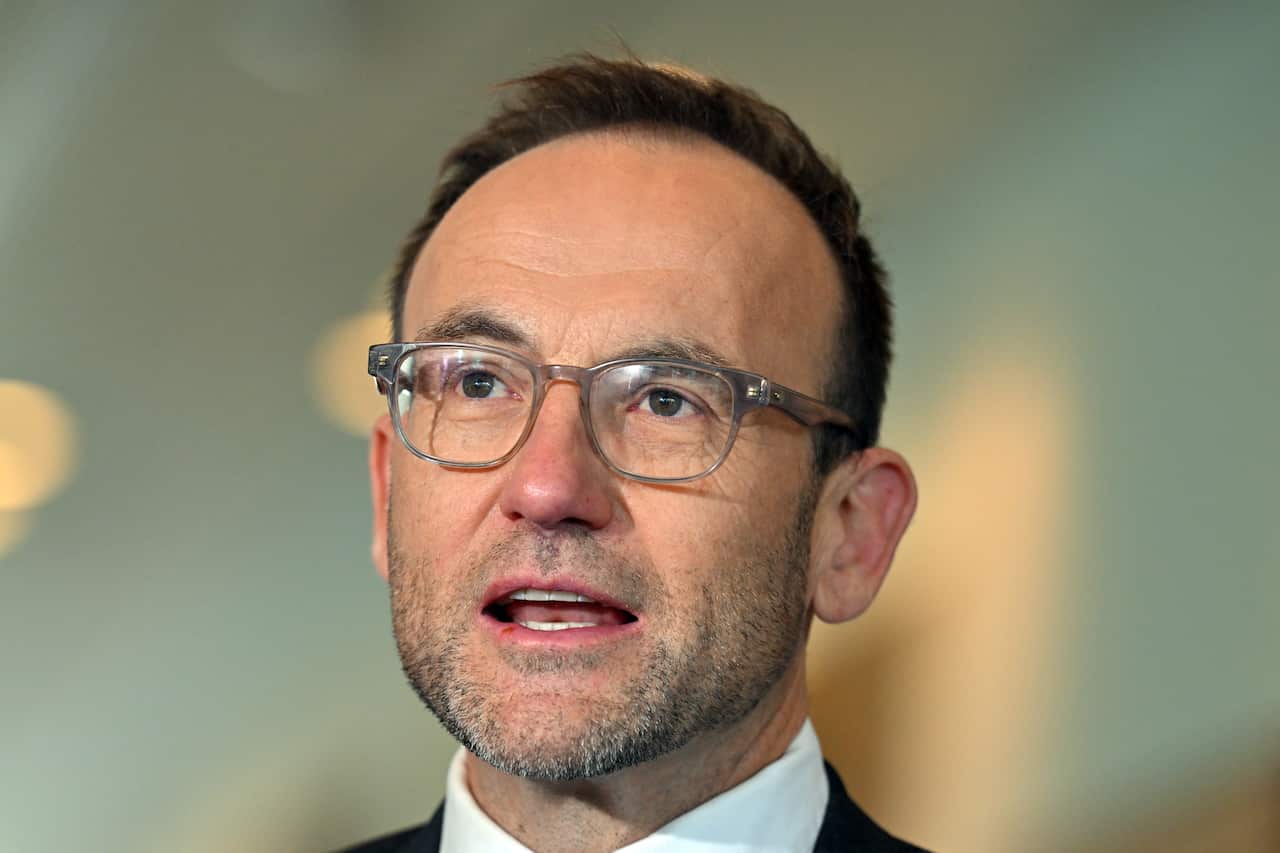 A close up of Adam Bandt wearing a suit and glasses speaking in a press conference