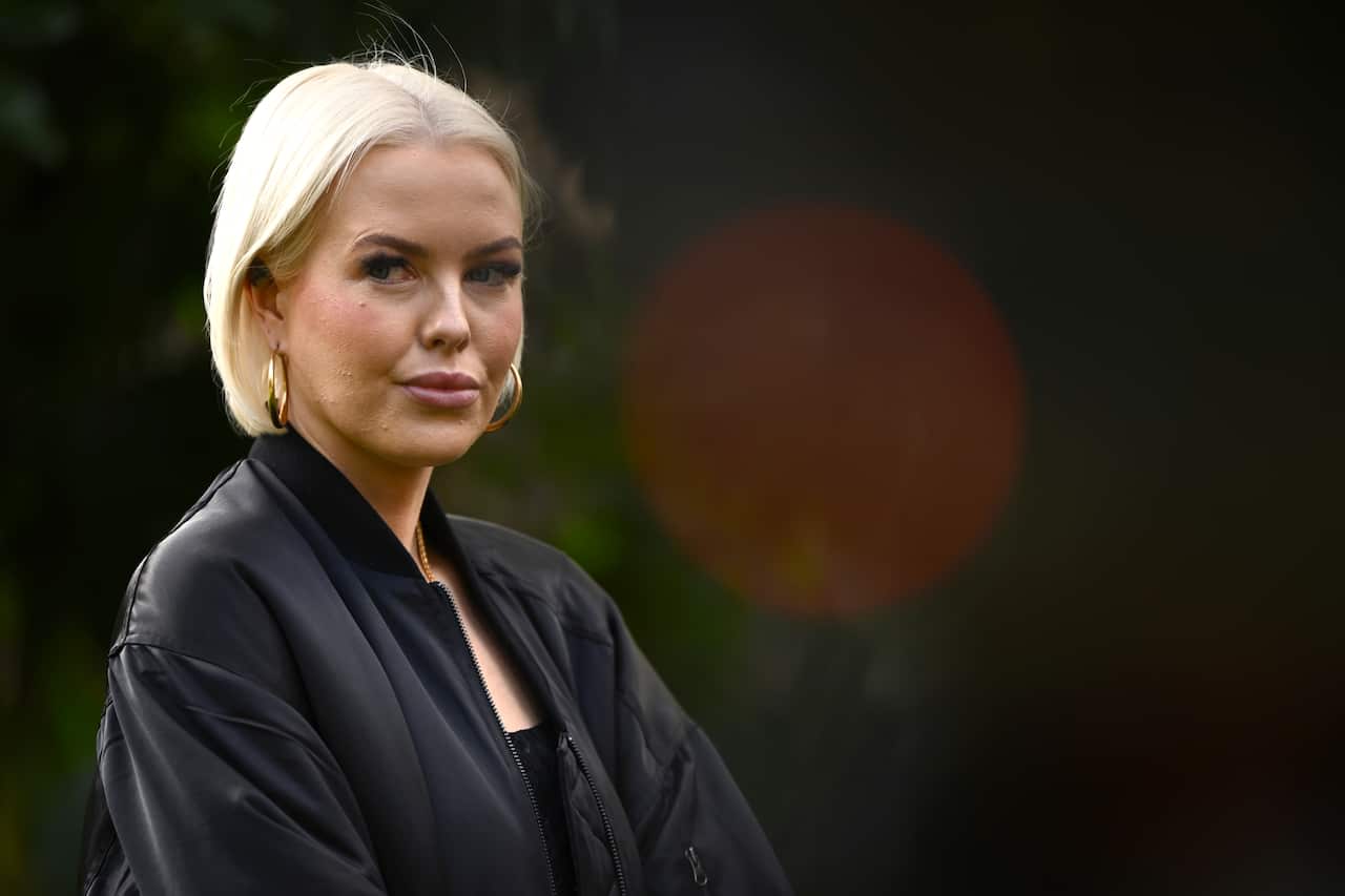 A woman in a black top and short blonde hair looks to her right with a serious expression on her face. 