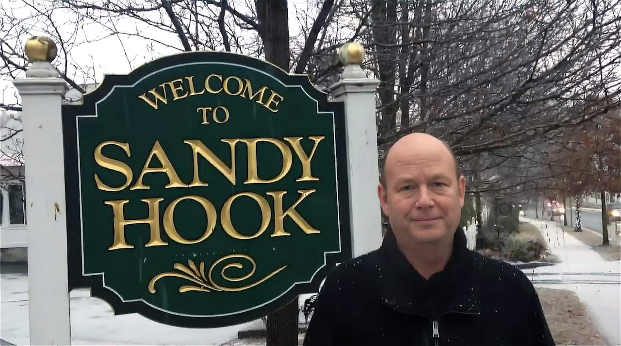 A man with a neutral expression stands by a sign that reads Welcome to Sandy Hook.