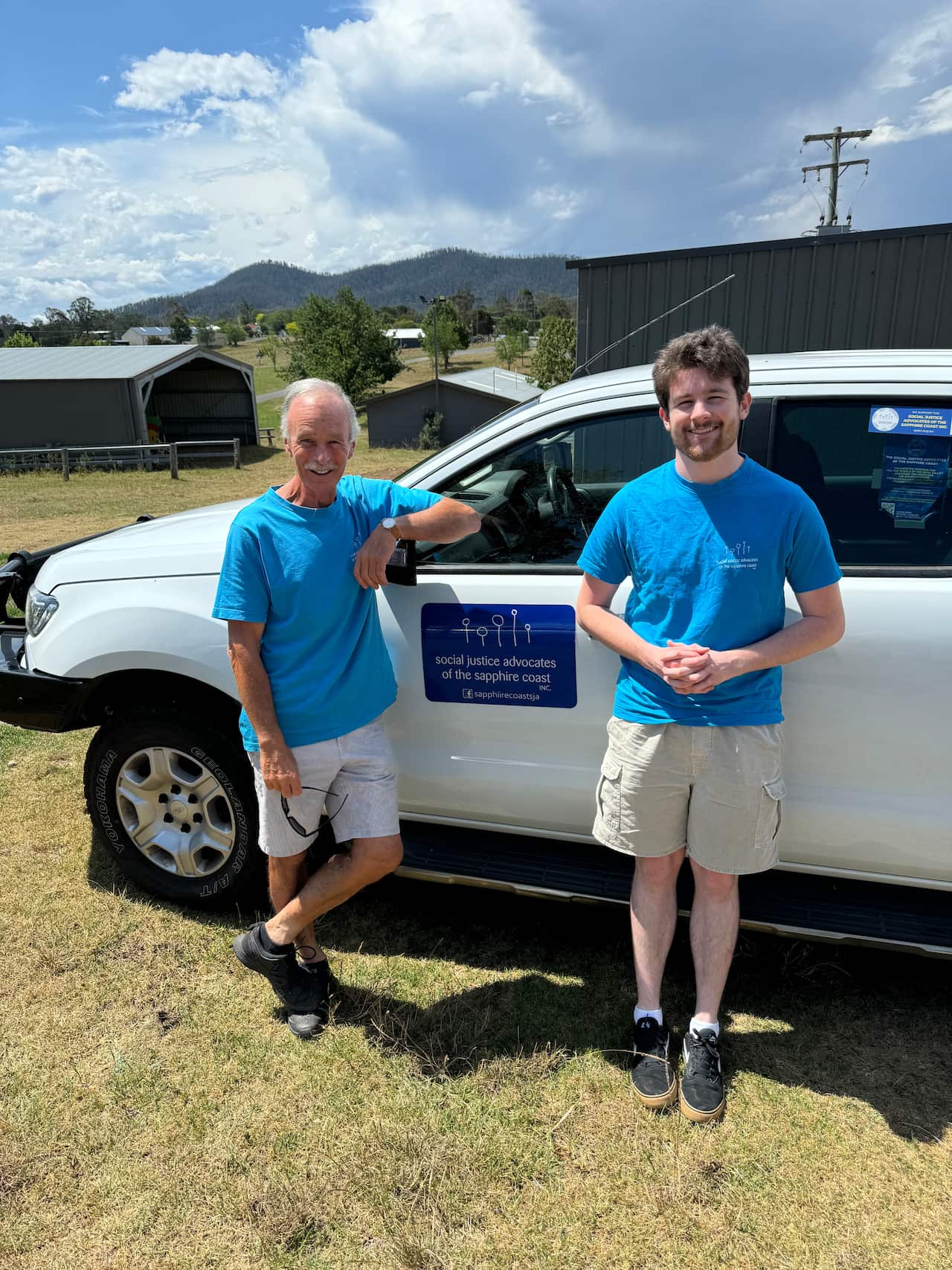 Two men in blue shirts smile as they stand in front of a ute.