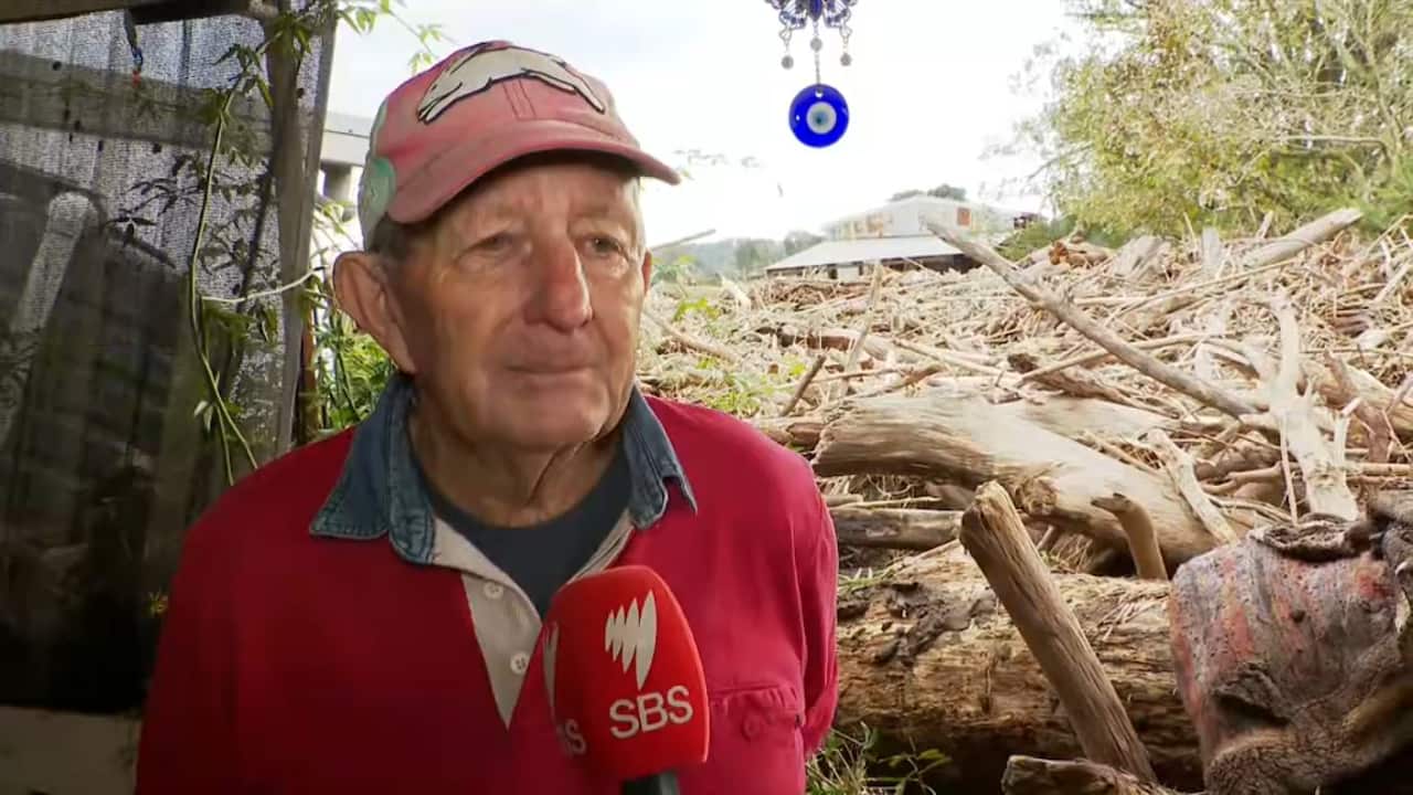 An older man wearing a red jumper and faded red sports cap. An SBS branded mic in front of him. He looks sad and exhausted. An expanse of debris stretches out behind him.