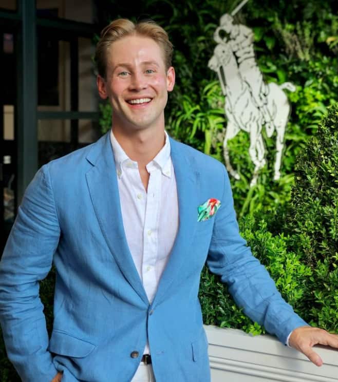 A man in a blue linen blazer, white shirt in front of a hedge.