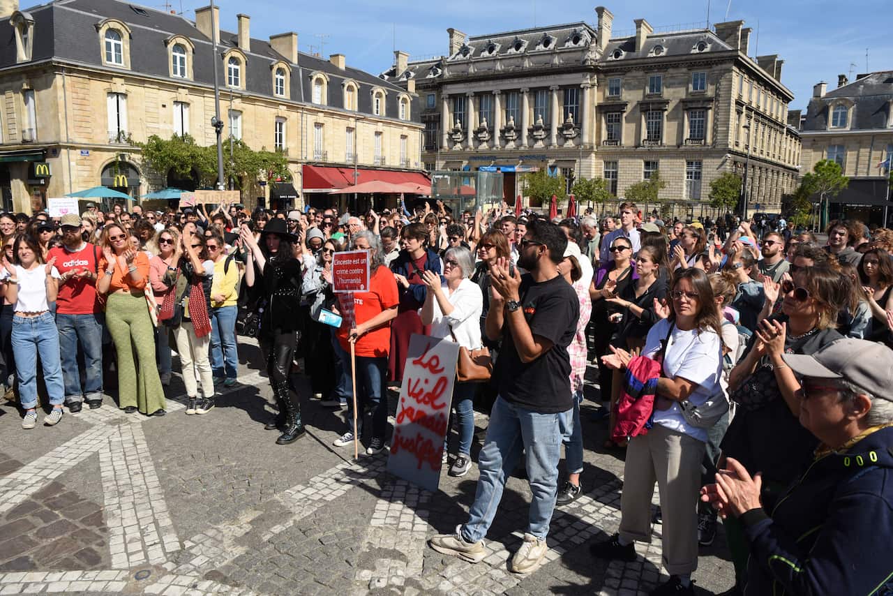 A crowd of demonstrators stand in a sunny boulevard in front of old buildings. 