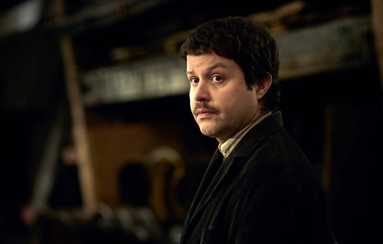 Young man with dark hair and moustache looks sideways.