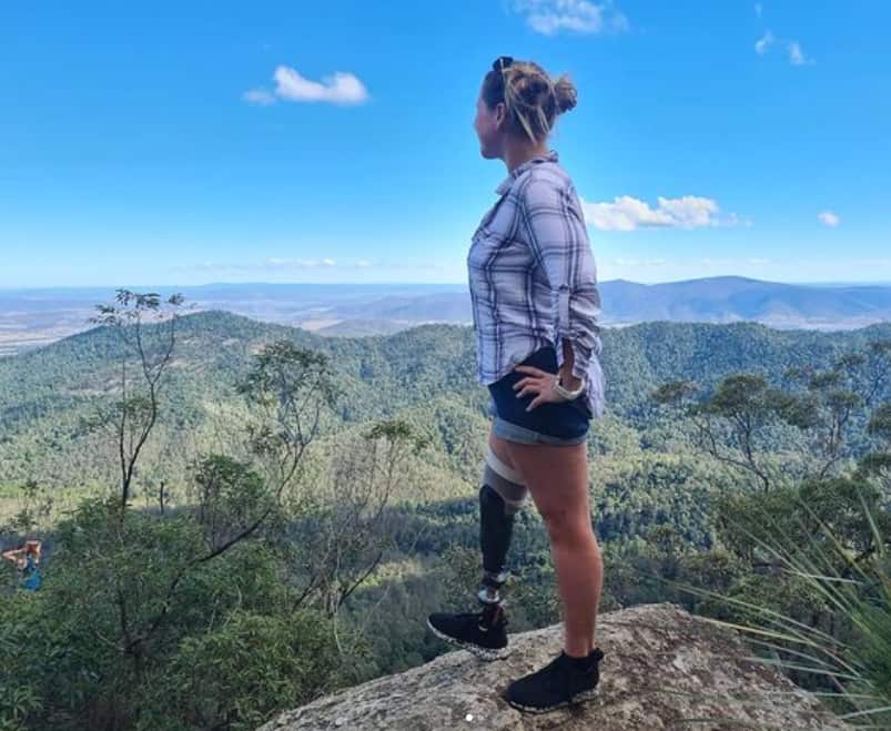 A woman with a prosthetic stands on a rock and looks out over the bush.