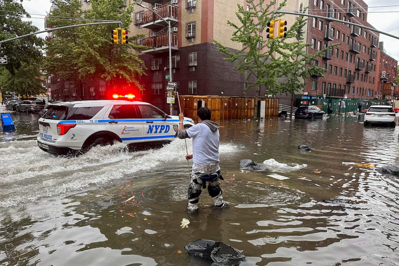 A man standing on a flooded street attempting to unblock a drain as a police car drives past.