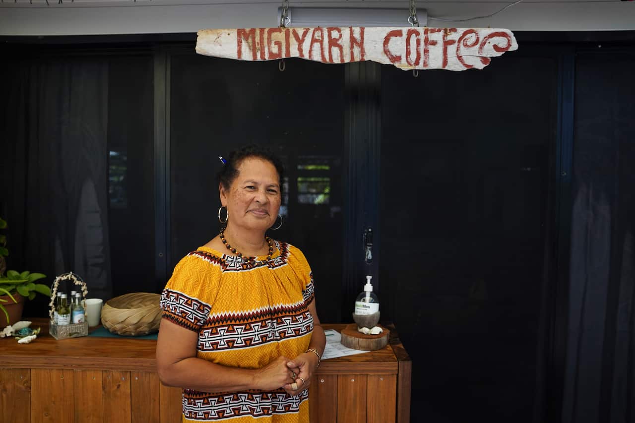 A woman wearing a yellow, patterned dress stands in front of a cafe with a sign reading 'MigiYarn coffee'.