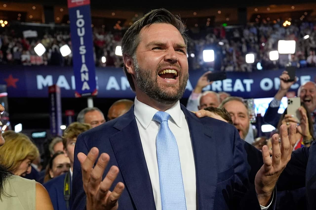 JD Vance wearing a dark blue suit jacket and light blue tie stands with a gleeful expression on his face with a crowd behind him at the Republican National Convention.