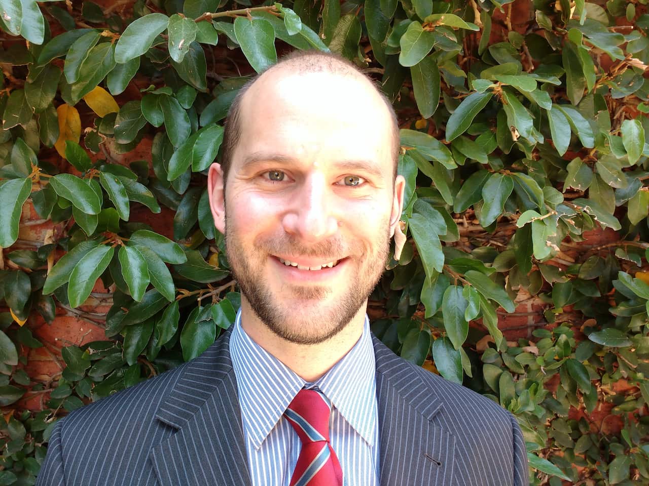 A smiling man with a beard and short hair, wearing a pinstriped suit and red tie, stands in front of a wall of green ivy and red brick.