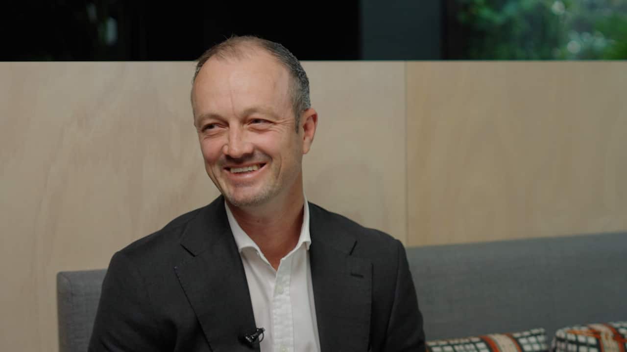A man in a dark suit and white shirt smiles off to the side while seated in a booth 