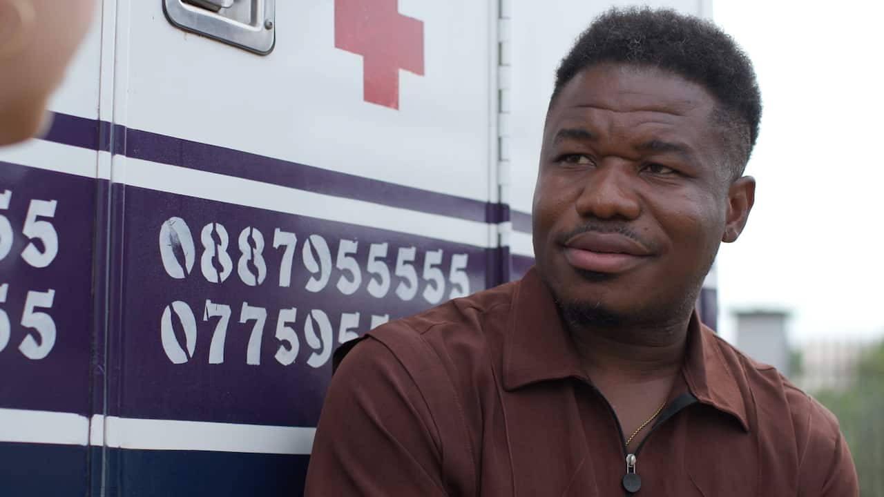 A man in a brown shirt sits beside an ambulance vehicle marked with a red cross symbol and emergency contact numbers.