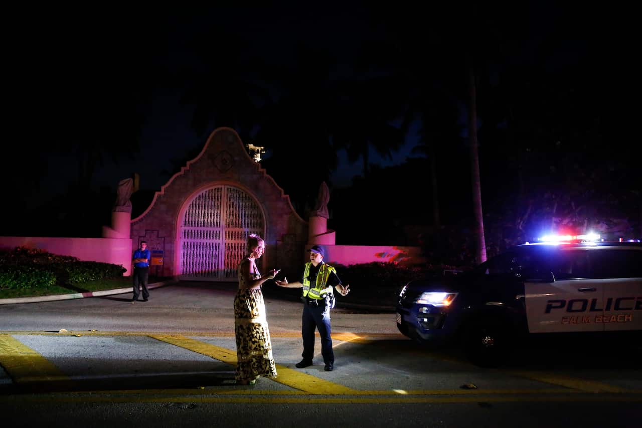 A woman speaking to a police officer who is standing in front of a police car with its lights on. It is dark outside.