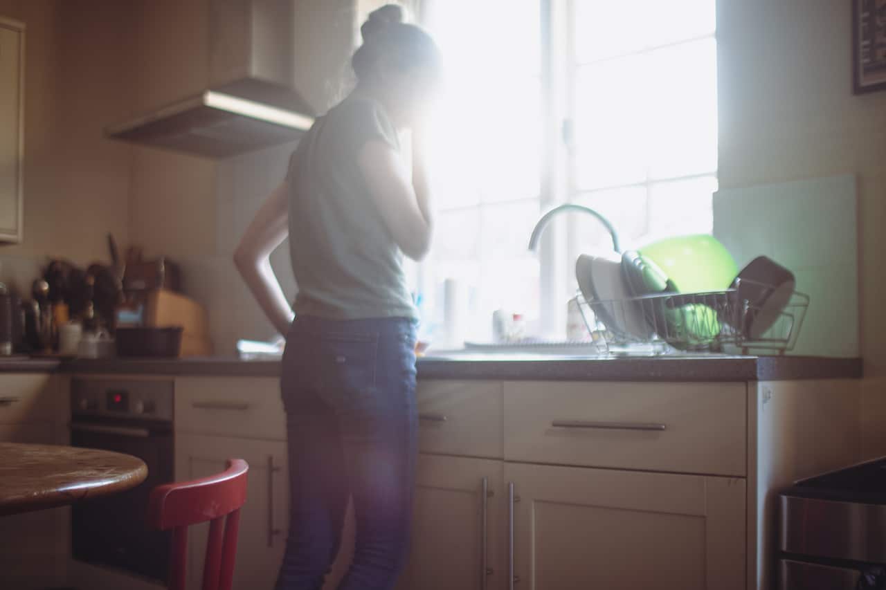 A woman, whose identity is blurred by sunlight coming through a kitchen window, stands at a sink.