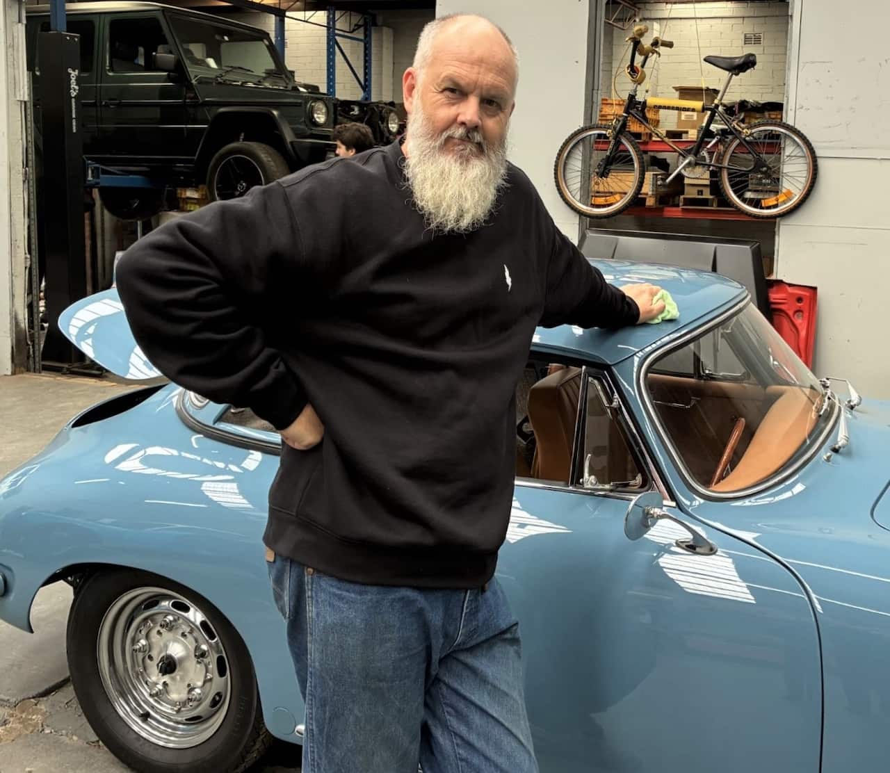 A man with a grey beard wearing a black sweater stands next to a blue vinage car in a workshop.
