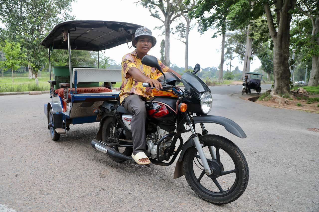 A man wearing grey pants, a yellow printed shirt, a grey bucket hat and white slip-on sandals posing for a photo on a tuk tuk 