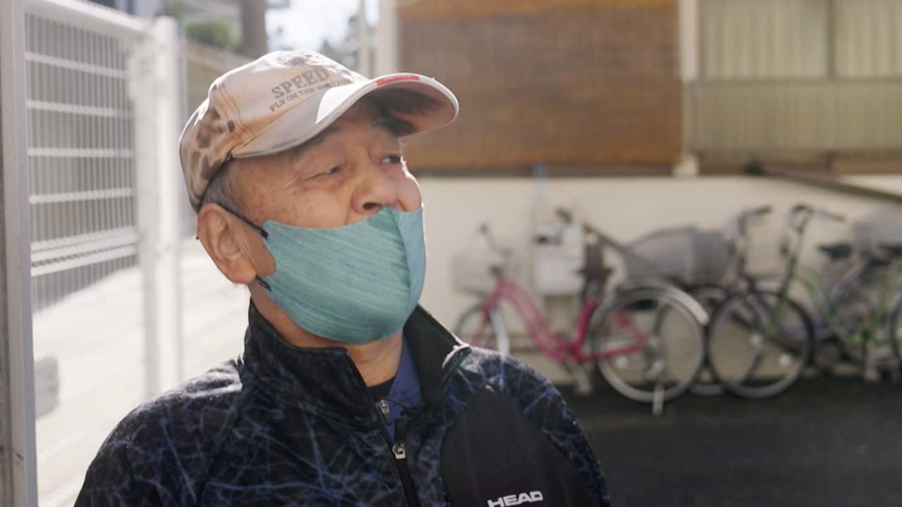 An elderly Japanese man wearing a beige baseball cap and a black jacket speaking with a blue medical mask covering the lower half of his face