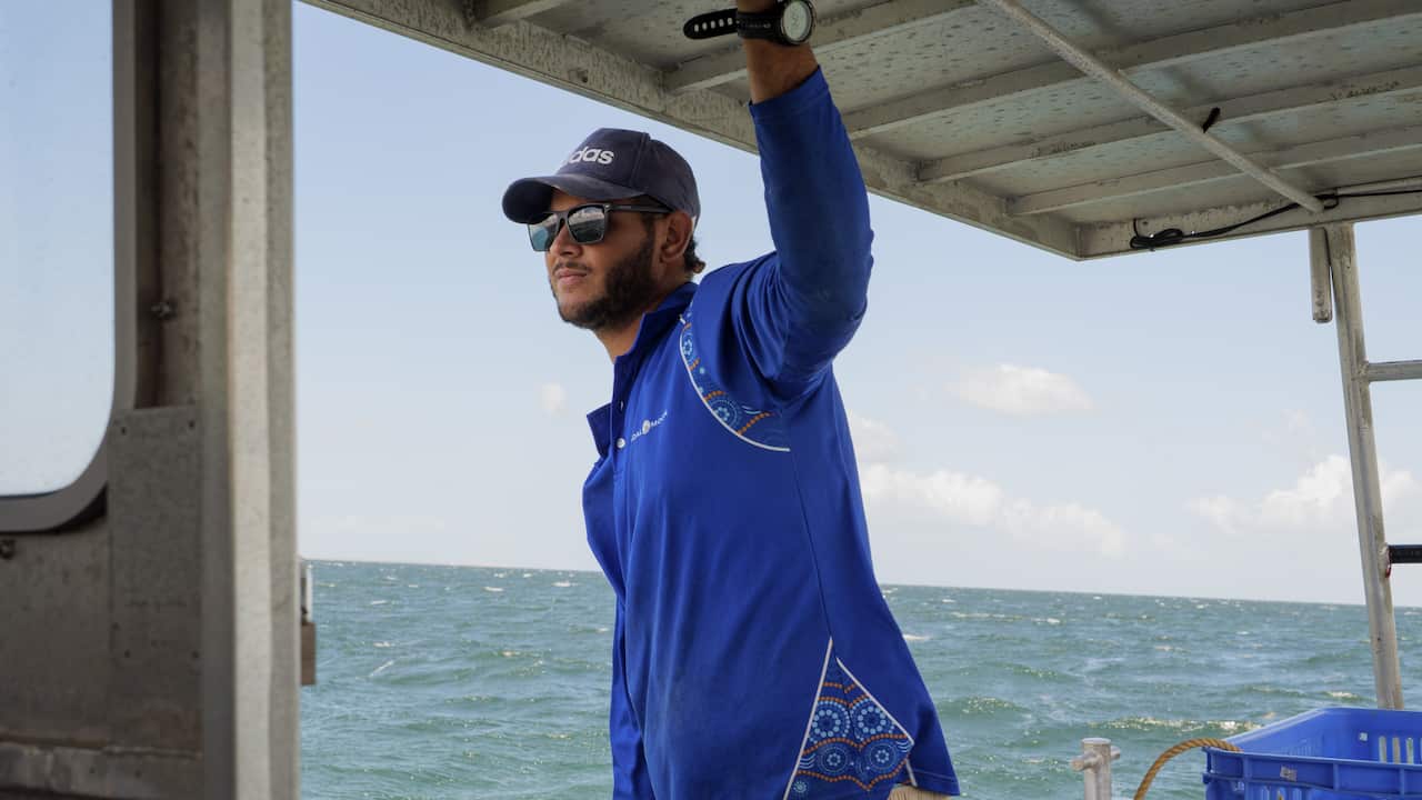 Shai, a young diver with Tidal Moon, wears a blue shirt, black cap and sunglasses and looks out over the ocean while holding onto the side of a boat.