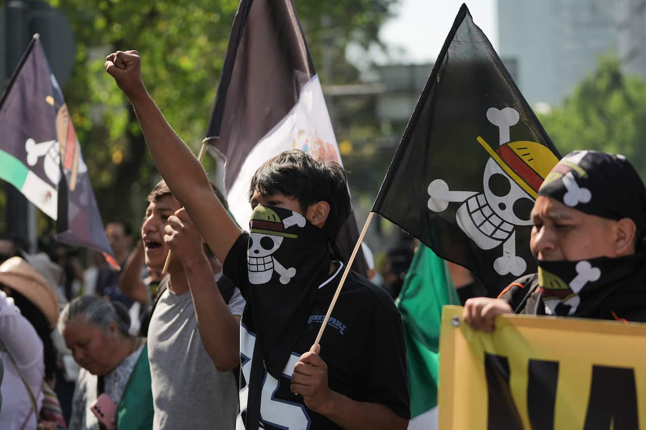 Protesters, some with their mouths covered with black scarves, with their hands raised, are holding black flags bearing skulls wearing yellow hats. 