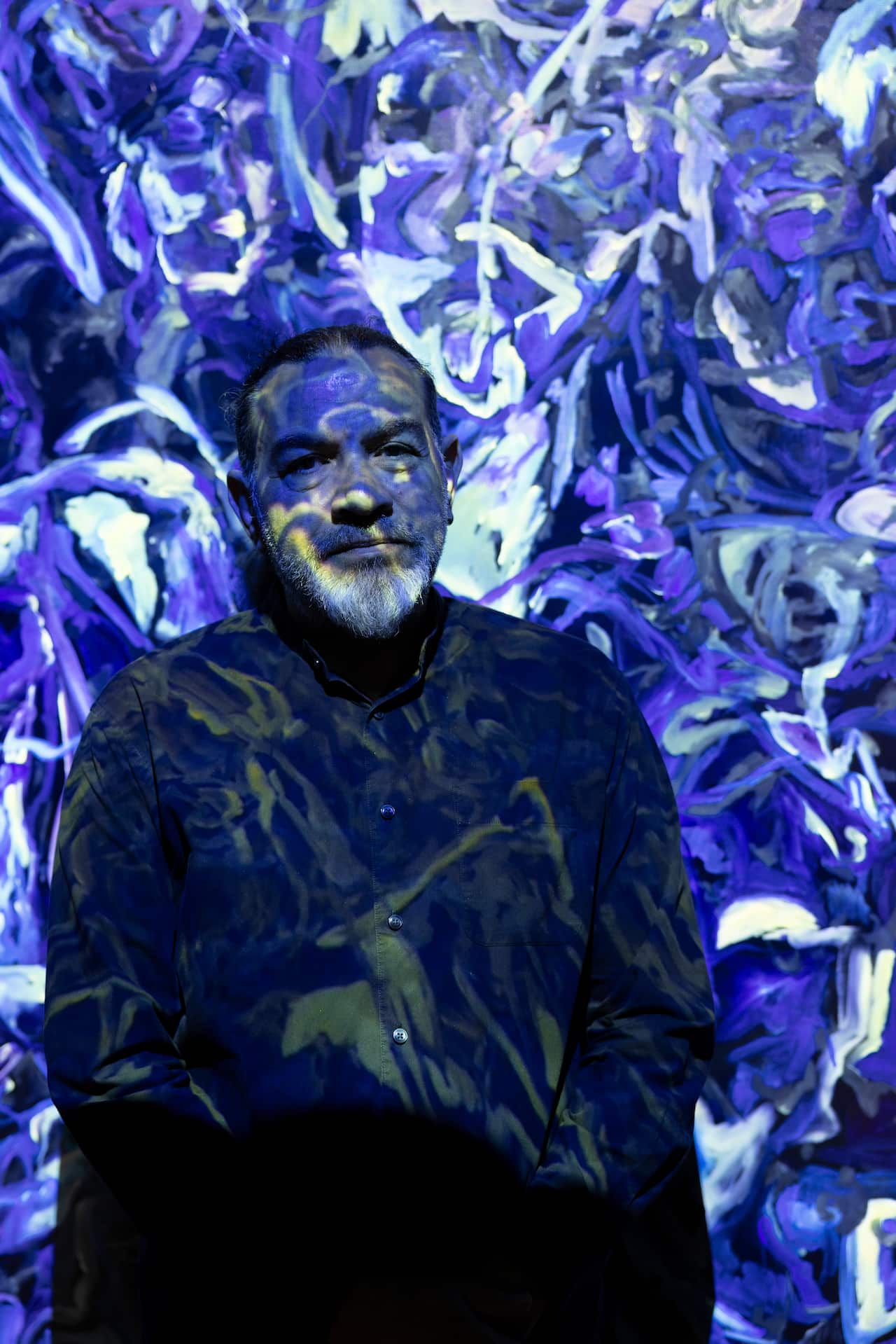 Khaled Sabsabi, a Lebanese Australian man with long hair, stands against a painted wall, with vivid purple and blue swirls are projected over him.