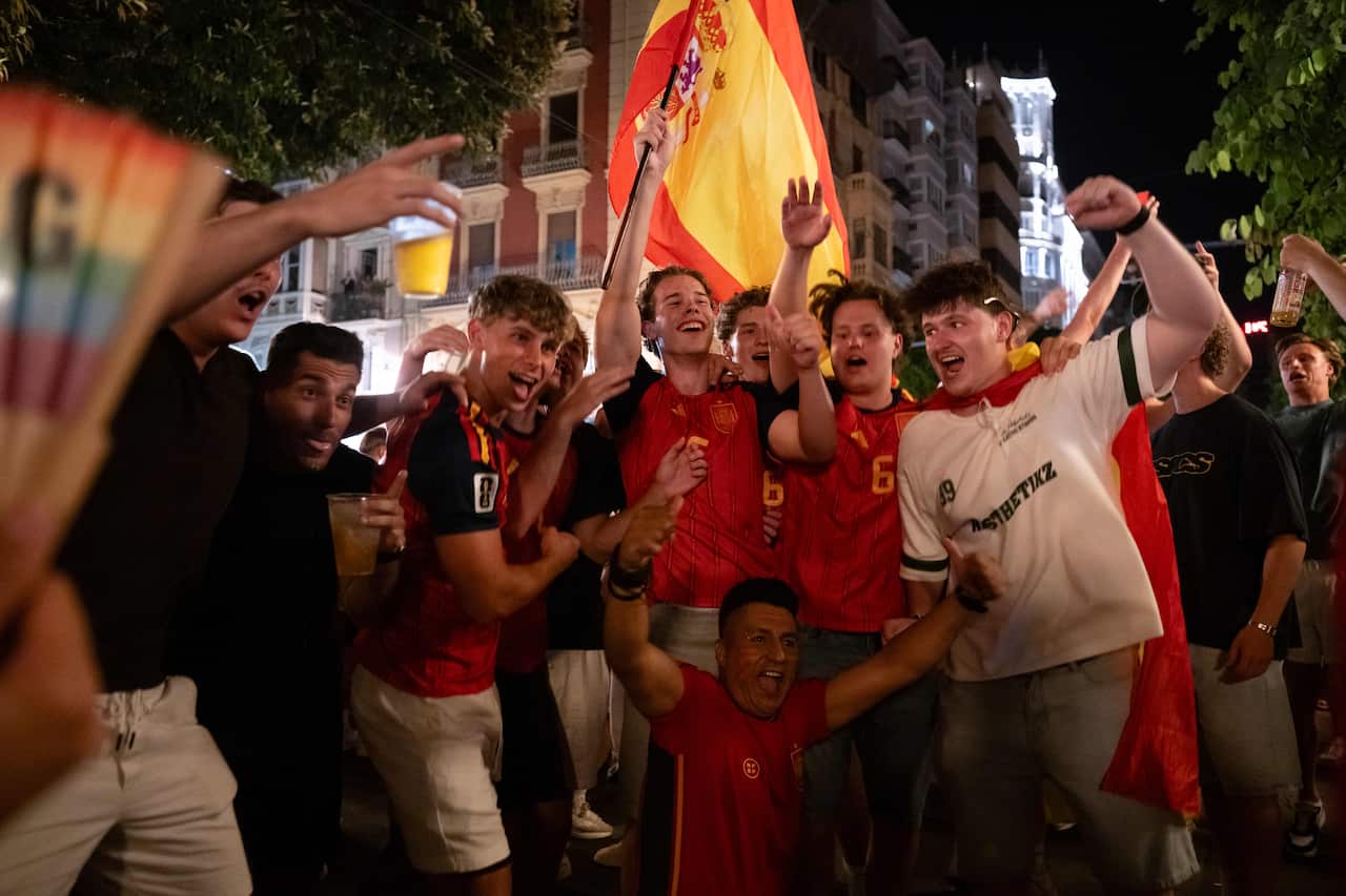 A crowd of happy fans celebrate Spain's win at the 2026 World Cup. They are dressed in Spanish red with fists raised. One carries a large Spanish flag.