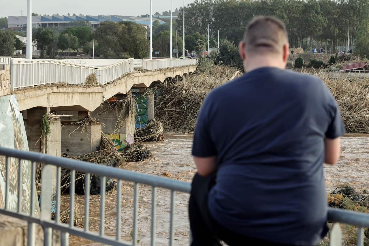 A man looks out at a brown river with a collapsed bridge
