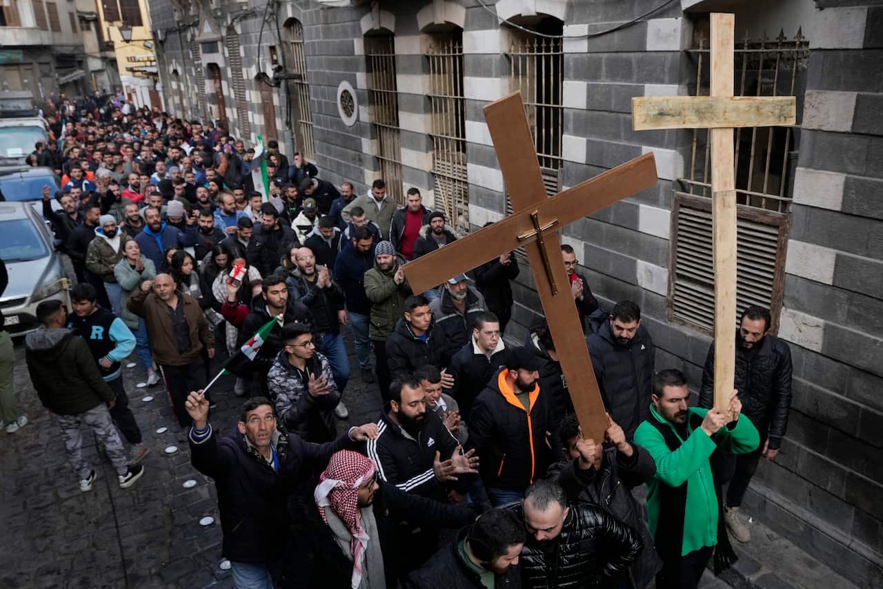 A large group of people march down a cobblestone street. Two people at the front of the march carry large wooden crucifixes. 
