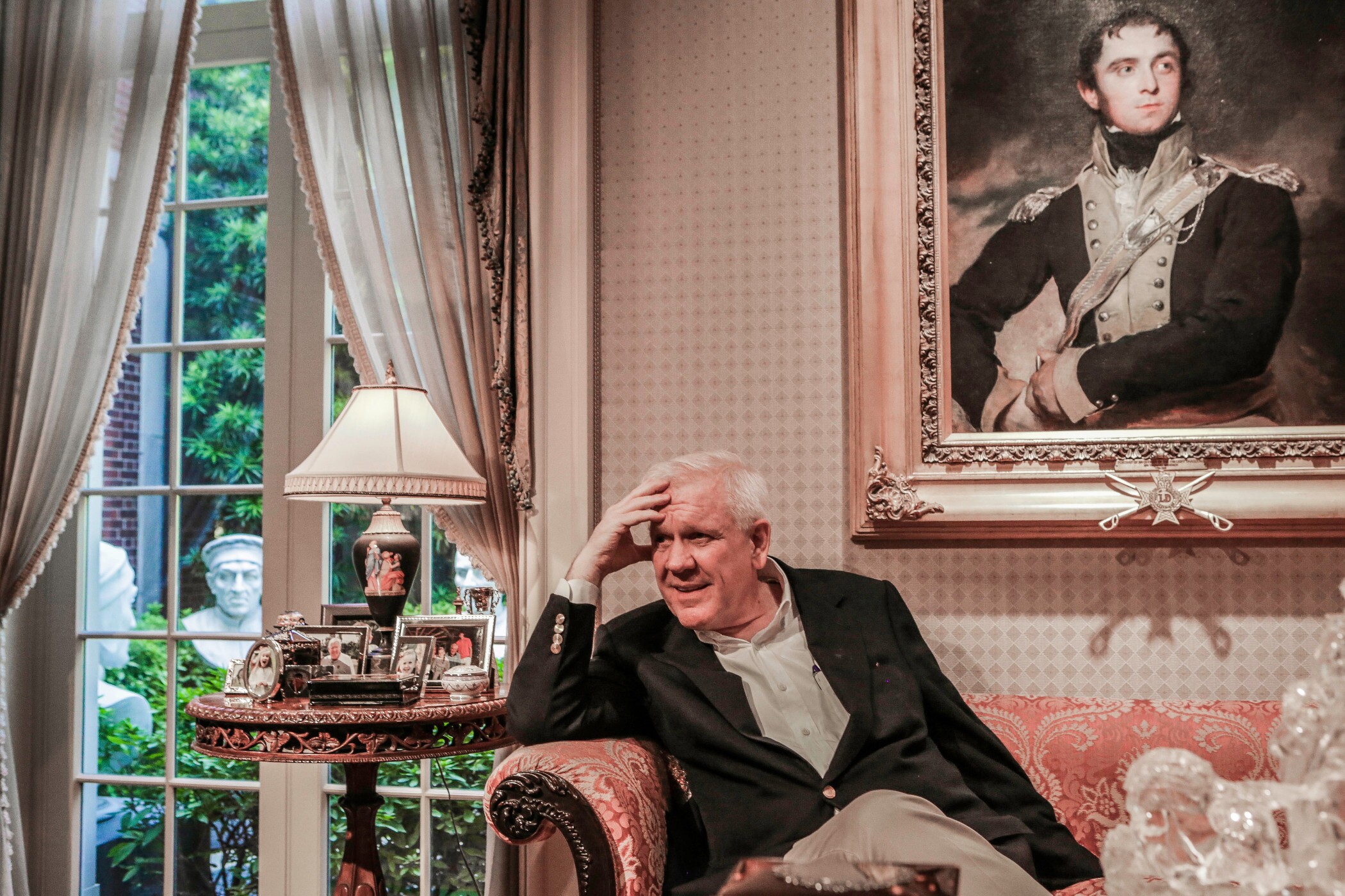Man with short, white hair in black blazer and white collared shirt sits on a pink salmon couch with his hand on his temple. 