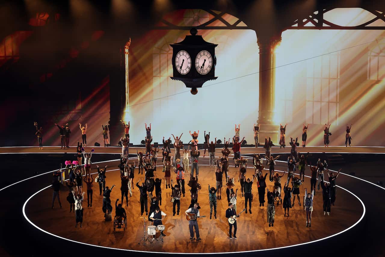 People dancing on stage. There is large old-fashioned four-sided clock suspended above them.