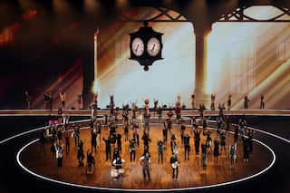 People dancing on stage. There is large old-fashioned four-sided clock suspended above them.