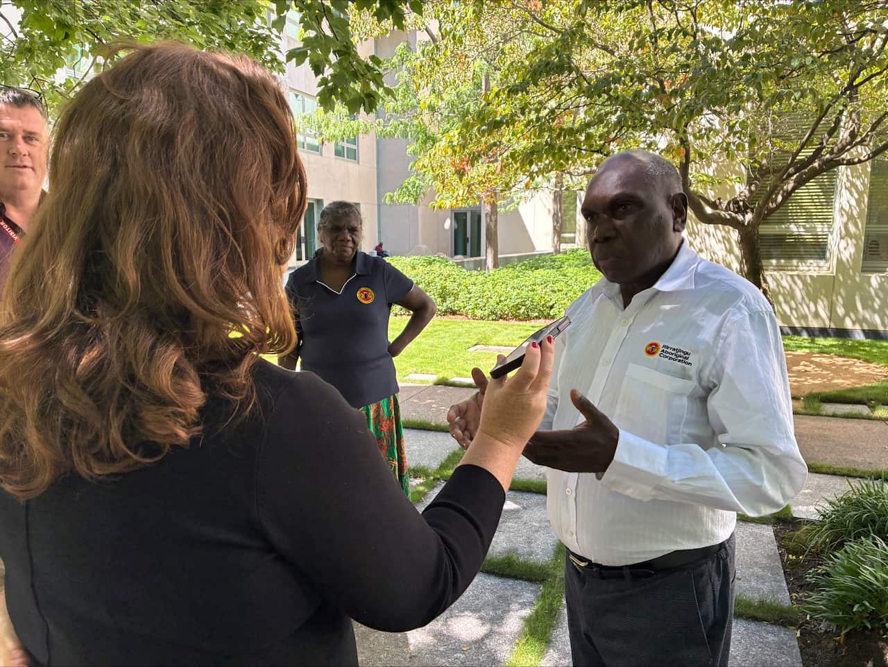 The back of a woman's head as she interview an Indigenous man in a white shirt and pants. The woman is holding a microphone.