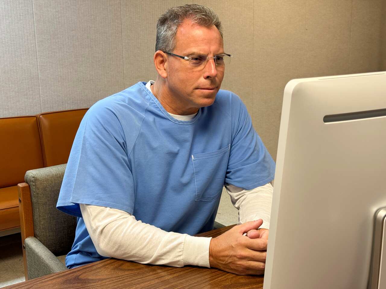 A man with short grey hair and wearing glasses and a blue short-sleeved smock over a long-sleeved white t-shirt. He is sitting at a chair at a desk and looking at a grey monitor.