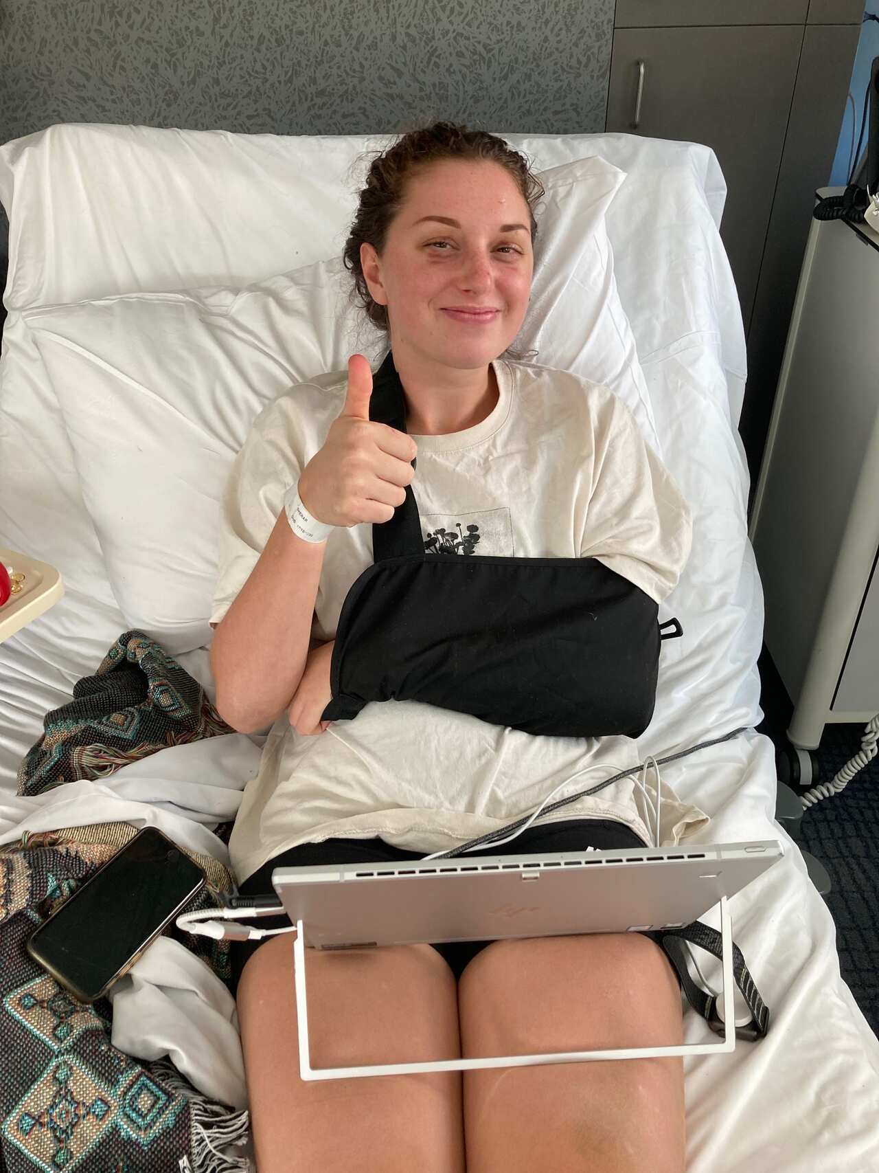 A woman with an arm in a sling sits in a hospital bed and smiles with her thumbs up.